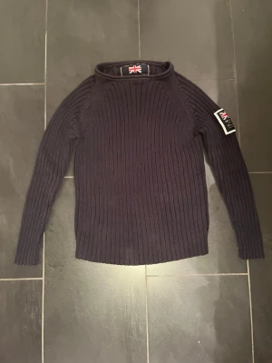 Mörkblå stickad tröja från Morris - Snygg mörkblå stickad tröja från Morris Ivy League med ribbstickat mönster och patch på ena ärmen. Tröjan har rund halsringning och är tillverkad i ett mjukt, värmande material. Perfekt för dig som gillar klassisk och stilren look.