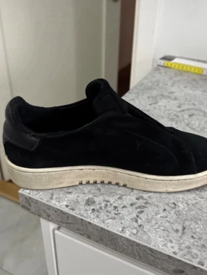 Svarta Axel Arigato laceless - Stilrena svarta Axel Arigato skor  från Axel Arigato med ovandel i sopigt mockaliknande material och vit sula. Skorna har en låg profil och diskret design utan snörning, vilket ger en clean och modern look. Perfekta för dig som gillar minimalistisk stil. Storlek 39 men passar 40