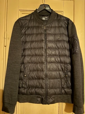 Svart pufferjacka från Polo Ralph Lauren - Snygg svart pufferjacka från Polo Ralph Lauren med quiltad front och ärmar i sweatshirtmaterial. Jackan har dragkedja, ribbade muddar och två fickor med knappstängning. Liten Polo-logga på bröstet och en extra ficka på ärmen för cool detalj.Obs ! Liten lagning som knappt syns , se sista bild . Obs ! Stl L men passar mer en i stl M
