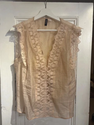 Beige blus med spets och knyt - Supersöt beige blus från H&M med djup v-ringning, spetsdetaljer längs kanterna och knyt i ryggen. Ärmlös modell i tunn, lätt bomull med romantisk känsla. Perfekt för dig som gillar en feminin och bohemisk stil.