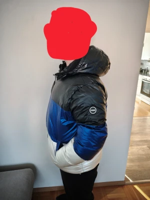 Blå, svart och vit pufferjacka OMBRE - Snygg pufferjacka från OMBRE i färgblock med svart upptill, blått i mitten och vitt nedtill. Jackan har huva, dragkedja framtill och två fickor. Perfekt för kalla dagar och ger en cool streetstil. Materialet är glansigt och vadderat för extra värme.