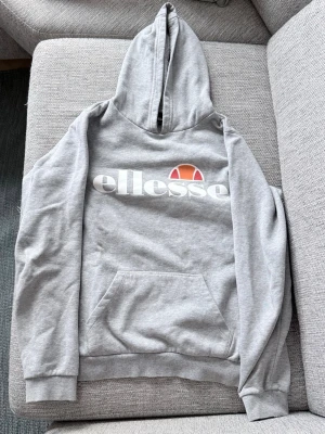 Grå Ellesse hoodie - Snygg grå hoodie från Ellesse med stor vit logga och orange detalj på bröstet. Klassisk känguruficka framtill, ribbade muddar och huva med dragsko. Tillverkad i mjuk bomullsblandning som är perfekt för chill dagar.