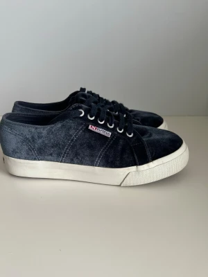 Mörkblå Superga sneakers i sammet - Snygga mörkblå sneakers från Superga i mjuk sammet med vita gummisulor och klassisk låg design. Skorna har svarta skosnören, metallöljetter och Superga-logga på sidan. Perfekta för dig som vill ha en stilren och trendig look.