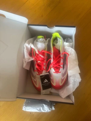 Adidas F50 Elite AG fotbollsskor - Säljer ett par Adidas F50 Elite AG fotbollsskor i vitt med neonrosa och gula detaljer samt svarta ränder. Skorna har en modern, strömlinjeformad design och är tillverkade i lätt syntetmaterial för bästa prestanda på konstgräs. Perfekta för dig som vill sticka ut på planen.