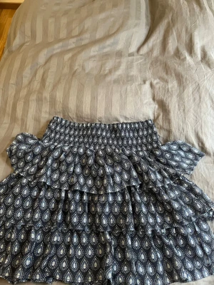 Mönstrad volangkjol från Lindex -  kjol från Lindex med mörkblå botten och vitt mönster. Kjolen har flera volanger och bred resår i midjan för skön passform. Perfekt för dig som gillar en lekfull och trendig stil.