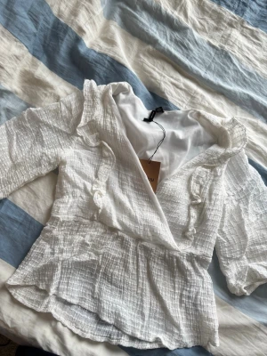 Vit omlottblus med volanger - Söt vit omlottblus från Bik Bok med volangdetaljer och knytband framtill. Blusen har ett krinklat tyg i bomullskänsla och långa ärmar. Perfekt för en trendig och avslappnad look.