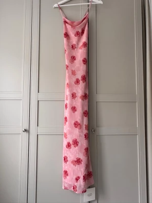Rosa blommig långklänning från ADOORE - Supersöt långklänning från Adoore i ljusrosa med röda blommor. Klänningen har tunna axelband, slimmad passform upptill och snörning i ryggen för extra detalj. Perfekt för sommarens alla tillfällen. Helt oanvänd prislappar kvar!! Modell Vienna maxi dress