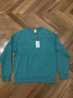 Turkos sweatshirt från Nike NOCTA - Snygg turkos sweatshirt från Nike NOCTA med klassisk rund halsringning och svart Nike-logga på bröstet. Tröjan har ribbade muddar vid ärmslut och nederkant samt NOCTA-detalj på ärmen. Perfekt för en avslappnad och trendig stil.