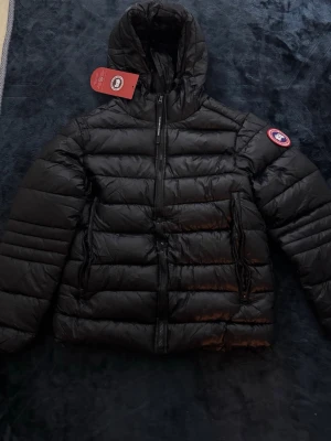 Jacka - Snygg svart dunjacka med huva och dragkedja framtill. Jackan har två stora fickor med dragkedja och klassisk logotyp på ärmen. Perfekt för kalla vinterdagar och riktigt varm tack vare dunfyllningen.