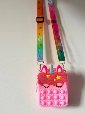 Rosa axelväska med enhörningsmotiv - Supercute rosa axelväska i silikon med pop-it detaljer och enhörningsmotiv. Väskan har färgglad justerbar axelrem med små söta mönster. Perfekt för dig som gillar unicorns och vill ha något lekfullt och unikt.