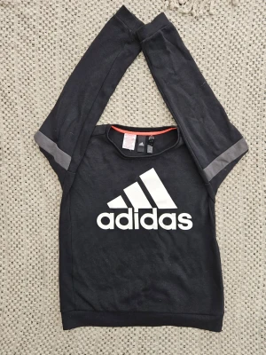 Svart Adidas sweatshirt med logga - Svart sweatshirt från Adidas med stor vit logga på bröstet och gråa detaljer på ärmarna. Tröjan har rund halsringning och långa ärmar. Perfekt för en sportig och avslappnad stil.