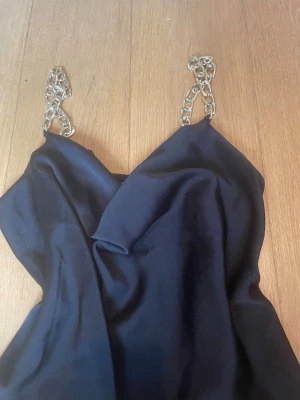 Svart topp med kedjeband - Snygg svart topp med bred urringning och guldfärgade kedjor som axelband. Toppen är ärmlös. Perfekt för dig som vill sticka ut med en unik detalj. Materialet känns mjukt och följsamt. Storlek är XS men känns mer som S
