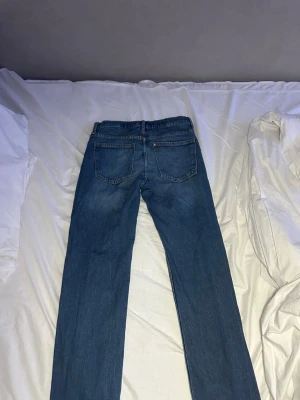 Blå raka jeans med fem fickor - Säljer ett par klassiska blå jeans med rak passform och fem fickor. Jeansen har normal midja, bälteshällor och stängs med knapp och dragkedja. Materialet är denim i bomull och färgen är en tidlös mellanblå nyans. Byxorna har storleken 158