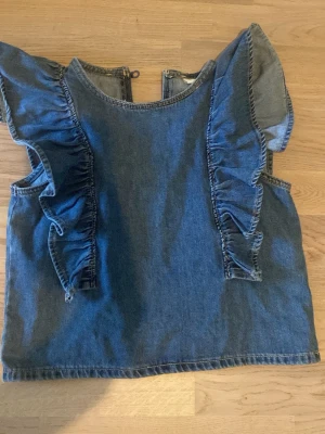 Blå jeanslinnetopp med volanger - Säljer en Snygg blå jeanslinnetopp med volangdetaljer längs axlarna och ärmlös design. Toppen har en rund halsringning fram och en v-ringad öppning bak. Perfekt för dig som gillar denim soppar och vill sticka ut lite extra. Den är sååå gullig! Jag har aldrig använt den så den är i väldigt  väldigt bra skick! Storlek 152 från Kappahl men passar nog någon som är lite längre också! Skriv om ni har några frågor!🩷