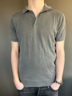 Grå pikétröja med krage - En stilren grå pikétröja med klassisk krage och två knappar framtill. Tröjan har korta ärmar och ribbade muddar vid ärmslut och nederkant. Perfekt för en avslappnad men ändå snygg look.  Modellen är 173cm 62kg. OBS! Pikén är sliten under ena armen!