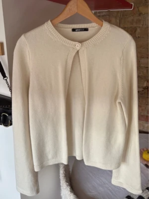 Krämvit kofta från Gina Tricot - Stilren krämvit kofta från Gina Tricot med rund halsringning och ett dekorativt knappfäste upptill. Koftan har långa ärmar och är tillverkad i ett mjukt, stickat material som passar perfekt till lager på lager. Enkel och clean design som är lätt att matcha.