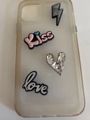 Två mobilskal med dekorationer -  iPhone 11. Säljer tre unika mobilskal med coola dekorationer. Ett skal är genomskinligt med motiv som blixt, 'Kiss', ett silverhjärta och 'love'. Det andra är lila med smiley i strass, bokstaven E, glass, 'hey!' och ett lila hjärta. Båda är i plast och passar dig som vill sticka ut. 1 för 30kr