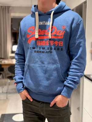 Blå Superdry hoodie med tryck - Snygg blå hoodie från Superdry med stort rött och vitt tryck på bröstet och japanska tecken. Hoodien har vit dragsko i huvan, känguruficka framtill och ribbade muddar vid ärmar och nederkant. Perfekt för en avslappnad streetwear-look.