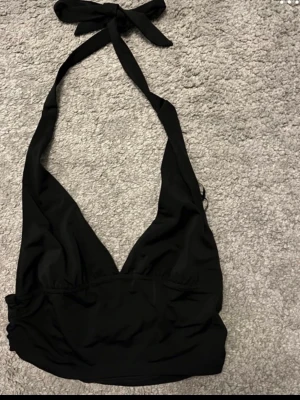 Svart bikiniöverdel med knyt - Snygg svart bikiniöverdel med halterneck och knyt i nacken. Djup v-ringning framtill och bredare band ger en trendig look. Perfekt för stranden eller poolen.