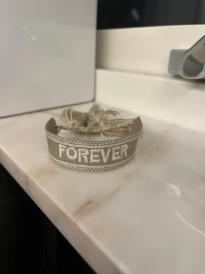 Beige tygarmband med FOREVER-text - Snyggt beige armband i tyg med broderad text 'FOREVER' i vitt. Armbandet har justerbara tofsar i ändarna för enkel passform och en stilren, avslappnad look. Perfekt accessoar för att lyfta din outfit.