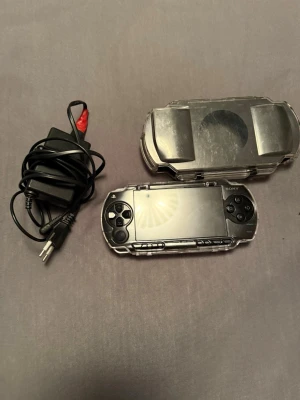 Sony PSP med laddare och skyddsfodral - Säljer en svart Sony PSP med tillhörande laddare och transparent skyddsfodral. Konsolen har klassisk oval form och knappar på sidorna av skärmen. Perfekt för portabel gaming och retrospel.