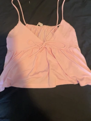 Ljusrosa omlott linnetopp med rosett - Supersöt ljusrosa linnetopp med tunna axelband och omlottdesign. Toppen har en dekorativ rosett framtill och är croppad för en trendig look. Perfekt för varma dagar och enkel att styla med jeans eller kjol.