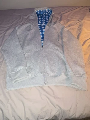 Grå hoodie med blåvitrutig huva - Säljer en grå zip war returning hoodie med dragsko och fickor framtill. Insidan av huvan har ett blåvitt rut pipeline-mönster som sticker ut och ger en unik look. Perfekt för dig som gillar street sop vibes och vill ha något bekvämt och stilrent. Lite oversized luva. Ett sample av Unbrandeds framtida hoodies