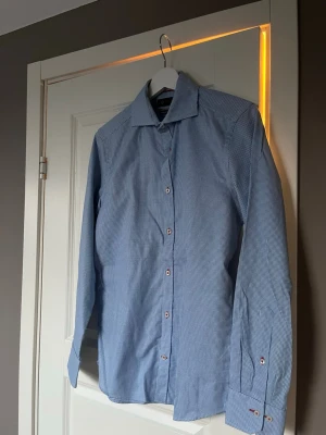Blå slim fit skjorta från Riley - Snygg blå skjorta från Riley i slim fit-modell. Skjortan har ett diskret rutmönster i vitt och blått, klassisk krage och långa ärmar med manschetter. Perfekt för dig som gillar en stilren och modern look.