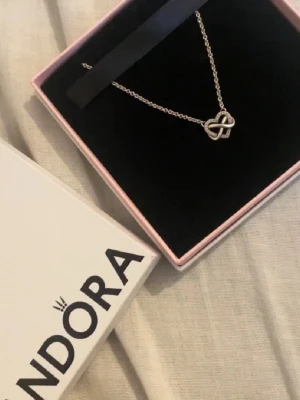 Pandora halsband med hjärthänge - Säljer ett stilrent silverfärgat halsband från Pandora med ett gnistrande hjärthänge i infinity-design. Hänget är dekorerat med små klara stenar som ger extra bling. Kedjan är tunn och elegant, perfekt för att lyfta vilken outfit som helst.
