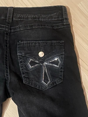 Gina tricot jeans med broderade fickor - Säljer ett par svarta jeans med raka ben och snygga broderade detaljer i silver på bakfickorna. Jeansen har låg midja och dekorativa knappar med logga. 