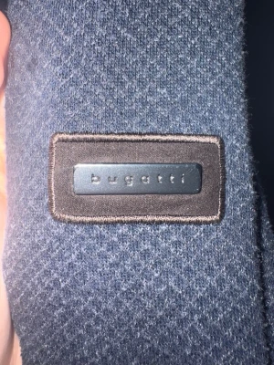 Mörkblå cardigan från Bugatti - Snygg mörkblå cardigan från Bugatti med ribbade muddar och krage. Jackan har dragkedja framtill och två fickor med dragkedja. Materialet är mjukt och har en lätt textur, vilket ger en modern och stilren look. Nypris 2000kr