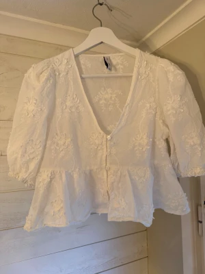 Vit blommig blus med volang - Superfin vit blus med broderade blommor och volang nertill. Djup V-ringning och knappar framtill. Puffiga trekvartsärmar ger en romantisk vibe. Perfekt till jeans eller kjol för en söt look. 