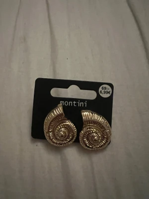 Guldiga snäckformade örhängen Montini - Säljer ett par guldiga örhängen från Montini med snäckformad design. Örhängena har en spiralformad struktur och glänsande finish som ger en cool och unik look. Perfekta för dig som vill sticka ut med accessoarerna.