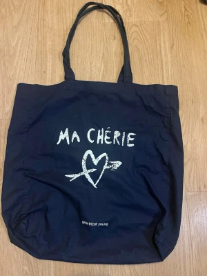 Svart tygkasse Ma Chérie Gina Tricot - Snygg svart shoppingväska i tyg från Gina Tricot Young med trycket 'MA CHÉRIE' och ett hjärta med pil på framsidan. Perfekt att slänga över axeln när du ska ut på stan. Rymlig och enkel att matcha med olika outfits.