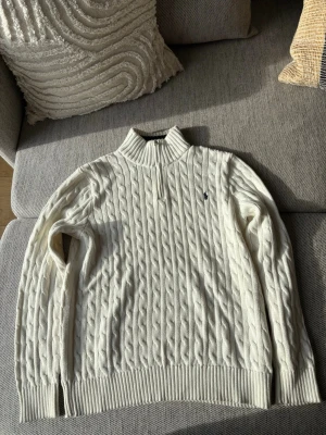 Vit kabelstickad tröja Polo Ralph Lauren - Kabelstickad vit tröja，halvzip och hög krage från Polo Ralph Lauren. Klassisk codetail med broderad logga på bröstet och ribbade muddar. Perfekt riktigt snygg till jeans eller chinos.