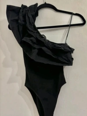 Svart body med volangdetalj - Säljer en svart body med snygga volanger över axlarna. Bodyn har en åtsittande passform och är tillverkad i ett mjukt, stretchigt material som känns bekvämt mot huden. Perfekt för att skapa en trendig look med lite extra edge.