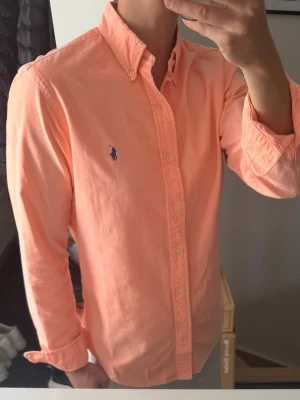 Persikofärgad skjorta från Ralph Lauren - Snygg persikofärgad skjorta från Ralph Lauren med klassisk button-down krage och broderad logga på bröstet. Skjortan har långa ärmar och knäppning framtill. Perfekt för en stilren och fräsch look. Den är ganska urtvättad, därav priset. men annars bra skick 