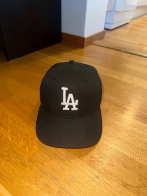 Svart LA-keps från New Era - Snygg svart keps från New Era med LA-logga broderad i vitt framtill. Klassisk basebollmodell, jag böjde skärmen lite grann dock. Perfekt för dig som gillar sportig och stilren streetwear.