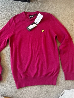 stickad tröja Lyle & Scott NY - Mörkrosa tröja från Lyle & Scott med rund hals och gul broderad logga på bröstet. Tröjan har långa ärmar och ribbade muddar vid ärmslut och nederkant. Tillverkad i mjukt material som känns skönt mot huden. FÄRG: burgundy som är en rosa röd ton 