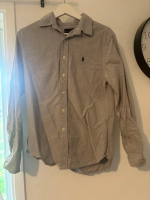 Grå skjorta från Ralph Lauren - Klassisk grå skjorta från Ralph Lauren med knappar framtill och broderad logga på bröstet. Skjortan har lång ärm och en tidlös design som passar till många olika stilar. Tillverkad i mjuk bomull för en bekväm känsla.