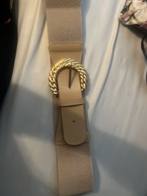 Beige bälte med guldspänne - Stilrent beige bälte med bred resår och ett snyggt, tvinnat guldspänne. Perfekt accessoar för att lyfta din outfit med en trendig touch. Bältet har detaljer i syntetmaterial och passar till både jeans och klänning.