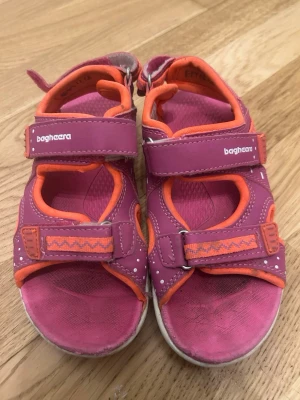 Rosa och orange sandaler från Bagheera - Snygga sandaler från Bagheera i rosa med orange detaljer och kardborreband. Sköna att ha på sig med mjuk innersula och justerbara remmar. Perfekta för sommaren och lätta att ta på och av. Tillverkade i syntetmaterial.