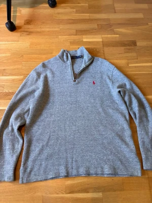 Grå halvzip tröja från Polo Ralph Lauren - Klassisk grå tröja med halv dragkedja från Polo Ralph Lauren. Tröjan har lång ärm, hög krage och den ikoniska röda logotypen broderad på bröstet. Tillverkad i mjukt material som känns skönt mot huden. Perfekt för en avslappnad men stilren look.