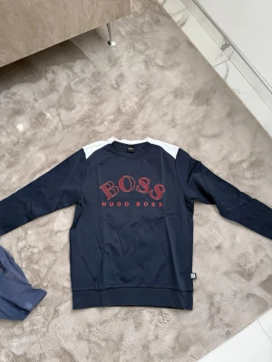 Mörkblå sweatshirt från Hugo Boss - Snygg mörkblå sweatshirt från Hugo Boss med röd logga och text på bröstet. Tröjan har vita detaljer vid axlarna och klassisk rund hals. Perfekt för en avslappnad och trendig stil.