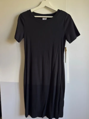 Svart ribbad klänning S 36/38 - En svart, ribbad klänning med kort ärm och rund halsringning. Klänningen har en enkel och stilren design som passar perfekt till många olika tillfällen. Materialet är mjukt och stretchigt för en bekväm passform.