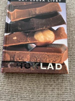 Det ljuva livet: Choklad - En bok med hårt omslag som handlar om choklad, fylld med inspirerande bilder på olika chokladbitar. Perfekt för dig som älskar sötsaker och vill läsa om chokladens värld. Omslaget är färgglatt med bruna och beige toner och stora bilder på choklad.