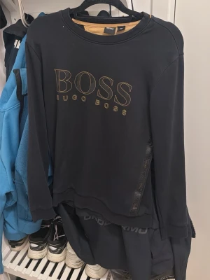 Svart sweatshirt från Hugo Boss - Svart sweatshirt från Hugo Boss med stort guldigt BOSS-tryck på bröstet. Tröjan har rund halsringning och långa ärmar. Tillverkad i mjukt material som känns skönt mot huden. Perfekt för en stilren och avslappnad look. Gammal modell från johnells