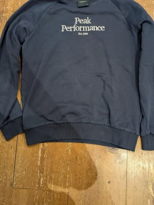 Mörkblå sweatshirt Peak Performance - Mörkblå sweatshirt från Peak Performance med broderad vit logga på bröstet. Klassisk rund halsringning och ribbade muddar vid ärmslut och nederkant. Perfekt för en avslappnad och stilren look.