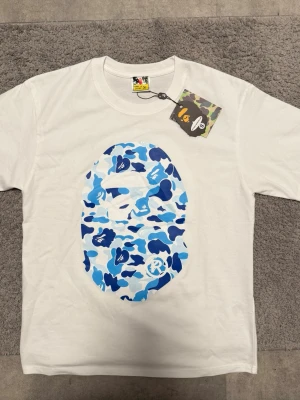 Vit BAPE t-shirt med blå camo - Säljer en vit t-shirt från BAPE med det ikoniska ape head-trycket i blått camo på både fram- och baksidan. T-shirten har rund hals och korta ärmar. Perfekt för dig som gillar streetwear och vill sticka ut med en unik design.