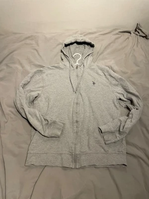 Grå hoodie från US Polo - Snygg grå hoodie med dragkedja från US Polo ASSN. Klassisk design med huva och dragsko, samt broderad logga på bröstet. Tillverkad i mjukt bomullsmaterial och har ribbade muddar vid ärmslut och nederkant.